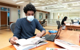 student using a laptop in the library while wearing a mask