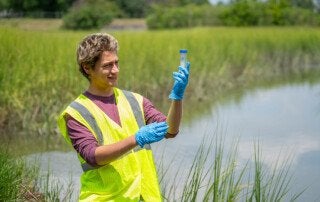 a man stands in marsh grass holding a test tube with water